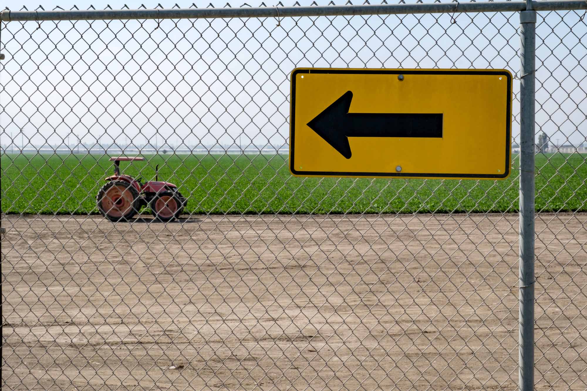 traffic sign pointing to the left hanging on a chaing-link fence, a field of crops and tractor beyond the fence