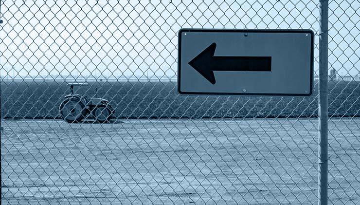 traffic sign pointing to the left hanging on a chaing-link fence, a field of crops and tractor beyond the fence